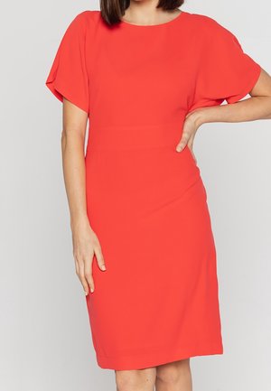 Femme portant une robe rouge vif, courte et à manches courtes, jusqu'aux genoux, debout avec une main sur la hanche et l'autre le long du corps.