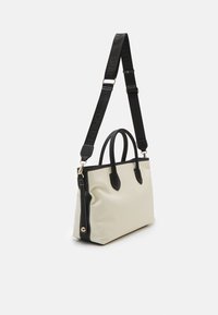 Beige tygväska med svarta läderdetaljer, dubbla runda handtag och en avtagbar svart rem med varumärkeslogga. Vita sömmar.