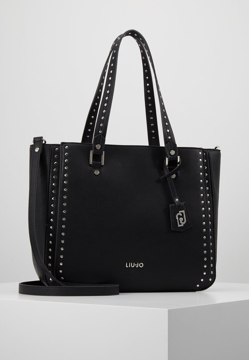 Sac fourre-tout en cuir noir avec accents cloutés, double poignées et une bandoulière amovible. Comprend une étiquette logo et une forme structurée.