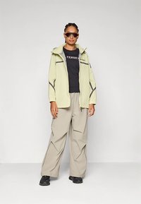 Φωτεινό πράσινο μπουφάν με φερμουάρ και γκρι λεπτομέρειες, μαύρο μπλουζάκι με λογότυπο, μπεζ παντελόνι με φαρδιά μπατζάκια και μαύρα παπούτσια· με αξεσουάρ γυαλιά ηλίου.