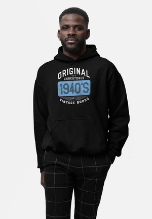 Homme portant un sweat à capuche noir avec le texte « Original and Unrestored 1940's Vintage Goods » et un pantalon noir à motif quadrillé blanc.