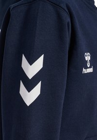 Mörkblå ärm med två vita chevronpilar och en delvis synlig vit kolibri-logotyp samt texten "hummel" på bröstområdet.