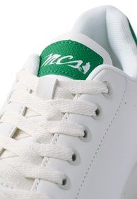 Zapatilla blanca de cuero con lengüeta textil verde, presenta cordones blancos y ojales de metal. Logotipo verde exhibido de manera prominente en la lengüeta.