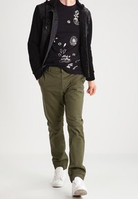 Persona vistiendo una camiseta gráfica negra, una chaqueta negra, pantalones verde oliva y zapatillas blancas caminando frente a un fondo simple.