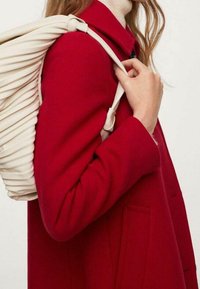 Personne portant un manteau rouge, portant un sac à dos plissé de couleur crème sur une épaule, la main tenant la sangle du sac.