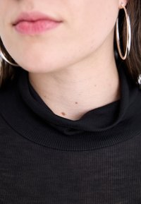 Černý žebrovaný rolák z jemné tkaniny, s volným designem na krku. Viditelné velké stříbrné kruhové náušnice, pleťová barva se pihami.