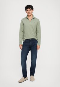 Ανοιχτό πράσινο μακρυμάνικο πουλόβερ με κλείσιμο προς τα επάνω, σε συνδυασμό με σκούρο μπλε τζιν και μπεζ loafers. Το μοντέλο στέκεται σε ουδέτερο φόντο.