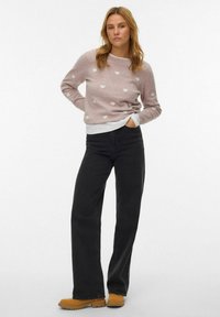 Roze trui met witte hartpatronen, laagjes over een wit shirt, gecombineerd met zwarte wijde jeans en bruine laarzen. Effen achtergrond.