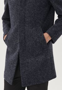 Cappotto in lana blu scuro con un motivo testurizzato, tasche laterali e un bottone frontale. Fittato, con una linea dritta e dettagli minimi.