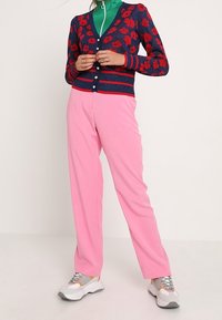 Kvinna i rosa byxor, vita sneakers, en grön zip-up-tröja och en marinblå kofta med röd blomsterdesign, ståendes med händerna hopknäppta.