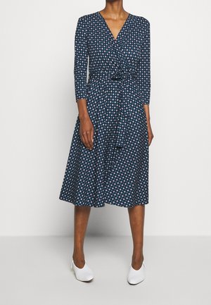 Kobieta w granatowej, midi sukience kopertowej z geometrycznym wzorem diamentów i rękawami do łokci, w połączeniu z białymi, spiczastymi butami.