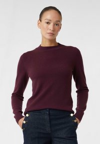 Vrouw met handen in haar zakken, gekleed in een fitted paarse, lange mouw trui en donkere, high-waisted broeken met gouden knopen aan de zijkanten.