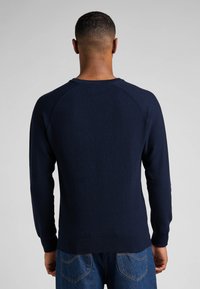 Pull en maille bleu marine avec un motif texturé, col rond et manches longues. Présente un ourlet et des poignets côtelés. Porté par-dessus un jean en denim.