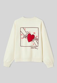 Sweatshirt de couleur crème avec un cœur rouge brodé entouré de mains, avec un contour graphique et une signature en bas.