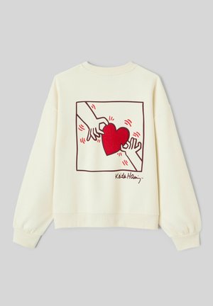 Sweatshirt de couleur crème avec un cœur rouge brodé entouré de mains, avec un contour graphique et une signature en bas.