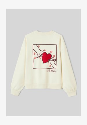 Sweatshirt de couleur crème avec un cœur rouge brodé entouré de mains, avec un contour graphique et une signature en bas.