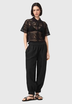 Kobieta ubrana w czarny krótki top z szydełkowym wzorem na rękawach na czarnym staniku, luźne czarne spodnie ze ściągaczami przy kostkach i czarne sandały z paskami wokół kostek.