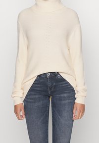 Donna che indossa un maglione a collo alto a coste color crema, infilato in un paio di jeans neri slim-fit sbiaditi, in piedi davanti a uno sfondo semplice.