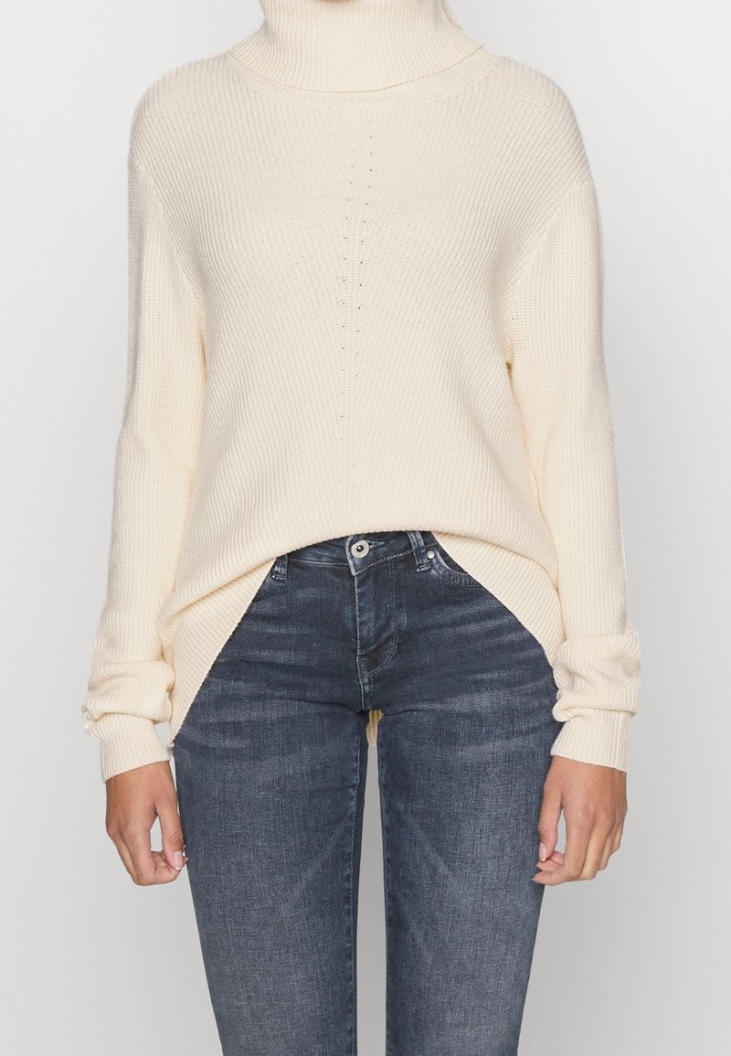 Donna che indossa un maglione a collo alto a coste color crema, infilato in un paio di jeans neri slim-fit sbiaditi, in piedi davanti a uno sfondo semplice.