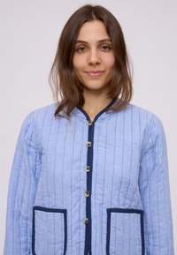 Giubbotto trapuntato azzurro chiaro con strisce verticali, rifiniture blu navy, cinque bottoni e due tasche quadrate. Presenta un collo rotondo e un tessuto testurizzato.