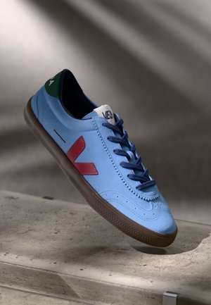 Baskets en suede bleu clair avec une semelle en caoutchouc marron, logo "V" rouge sur le côté, lacets bleu marine et accents verts près du talon.