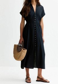 Abito nero a camicia realizzato in tessuto morbido, caratterizzato da una vestibilità comoda, maniche corte e una vita rifinita, abbinato a una borsa a tracolla intrecciata.