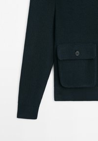 Donkerblauwe gebreide mouw van een trui met een voorzak met een klep en knoop op een witte achtergrond.