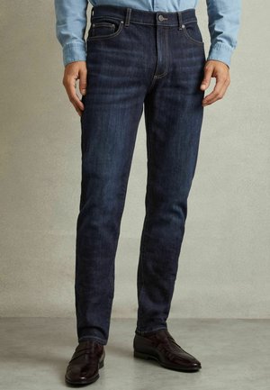Sötétkék farmernadrág, slim fit szabással, öt zsebbel, övfakkal és diszkrét varrási részletekkel. Sötétbarna mokaszinokkal párosítva.