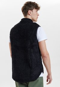 Schwarze, flauschige ärmellose Weste mit hohem Kragen, aus strukturiertem Material, getragen über einem weißen Hemd mit abgerundetem Saum.