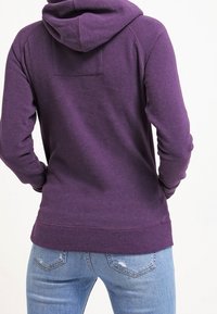 Sudadera morada hecha de una tela suave, con capucha ajustable y un bolsillo tipo canguro, estilizada con puños y dobladillo acanalados. Combinada con unos vaqueros azules.