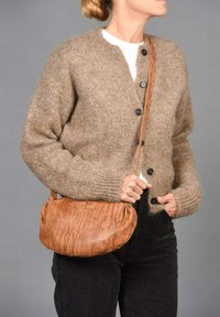 Cardigan de punto marrón con siete botones, llevado sobre una blusa blanca. Se sostiene de manera casual un bolso de hombro de cuero suave y arrugado en color marrón claro.