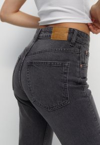 Jean en denim gris à taille haute, avec un patch en cuir brun au dos, des éléments en laiton cuivré et un style classique à cinq poches.