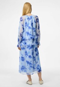 Prozračna plava cvjetna maksi haljina od prozirnog materijala, s dugim rukavima i nabranim slojevima. Sadrži velike uzorke plavih ruža i cvjetova.