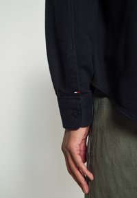 Primo piano della mano di una persona al suo fianco, che indossa una camicia a maniche lunghe scura con un piccolo logo rosso, bianco e nero sul polsino.