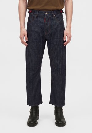 Homme portant un jean droit bleu foncé, des bottes noires à boucle et une chemise marron, debout devant un fond clair uni.