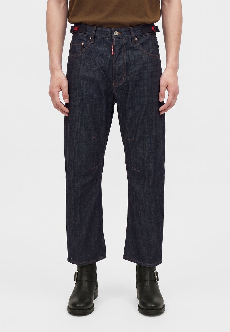 Homme portant un jean droit bleu foncé, des bottes noires à boucle et une chemise marron, debout devant un fond clair uni.
