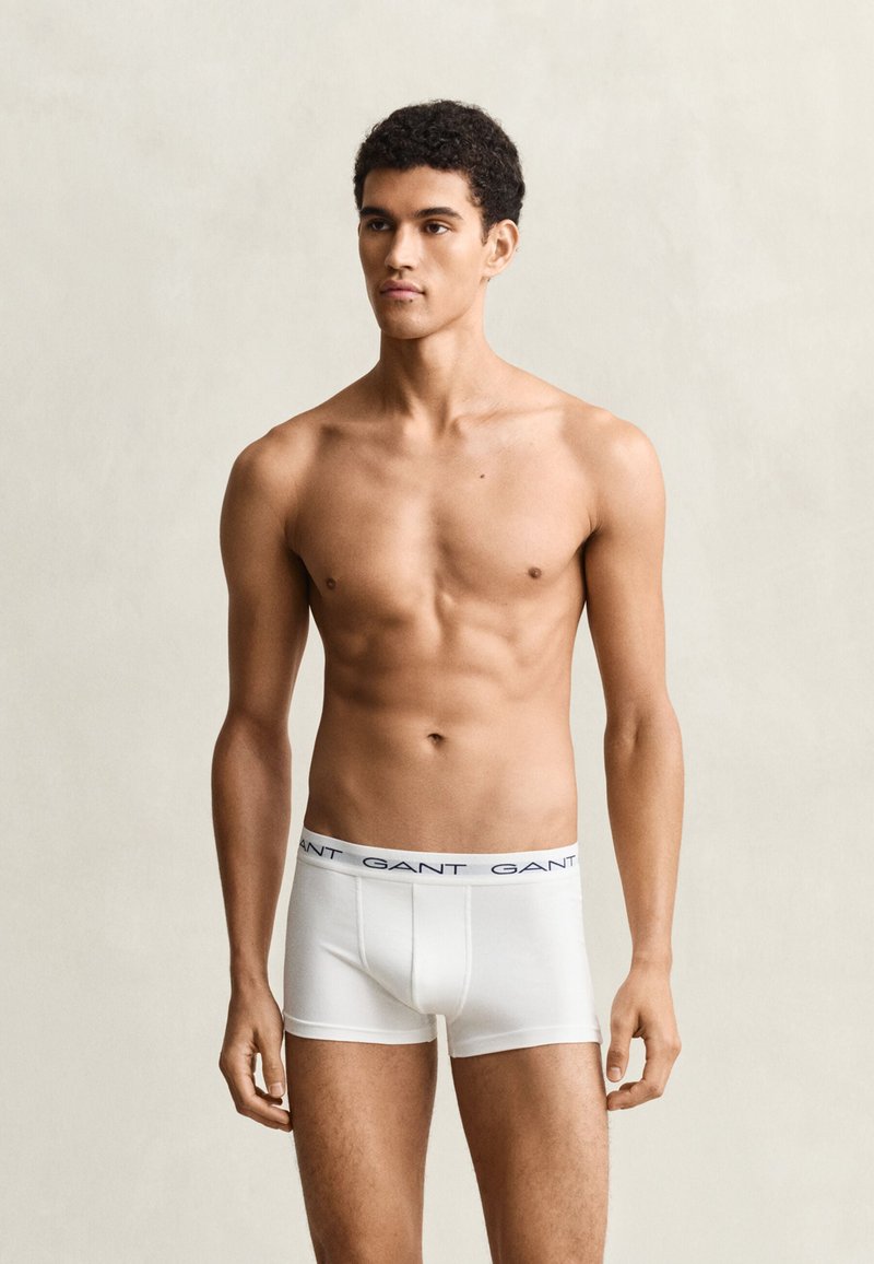 Jeune homme aux cheveux courts et bouclés, torse nu, portant des boxers blancs GANT sur un fond clair uni.