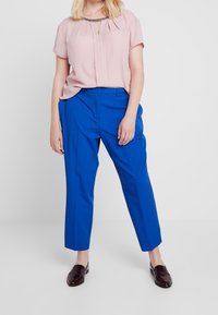 Ljusrosa blus med korta ärmar och plissédetalj, i kombination med ljusblå skräddarsydda byxor och mörkbruna loafers.