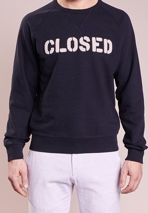 Mężczyzna w czarnej bluzie z napisem „CLOSED” białymi literami, w połączeniu z jasnymi spodniami.