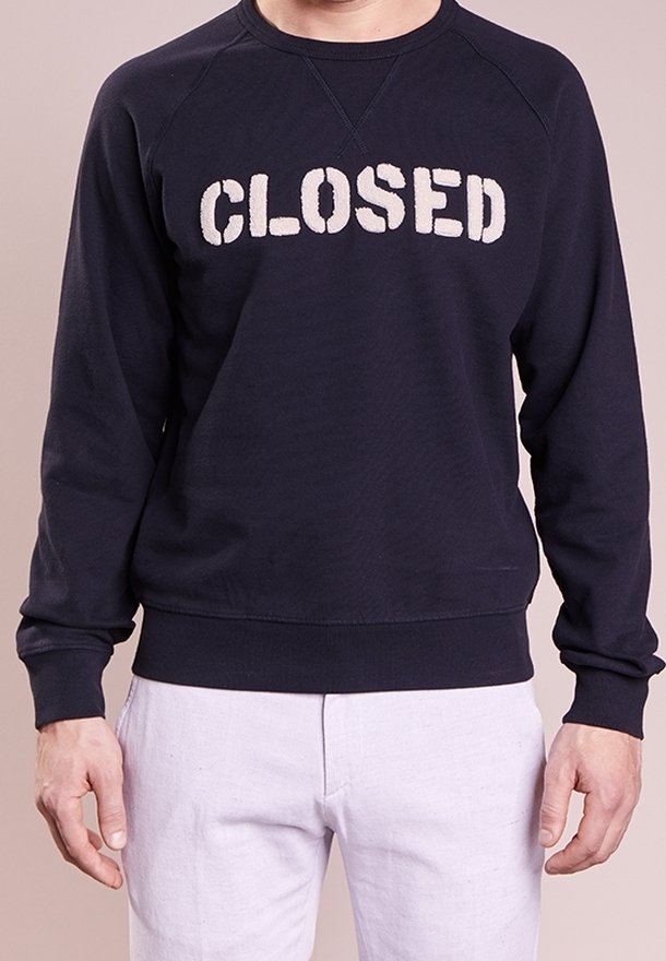Homme portant un sweat-shirt noir avec le mot « CLOSED » en lettres blanches, associé à un pantalon clair.