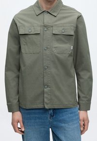 Homme portant une veste boutonnée vert olive avec deux poches poitrine et un jean bleu, debout devant un fond blanc uni.