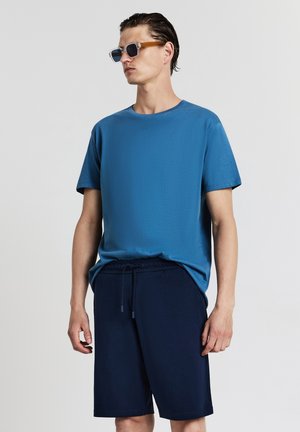 Blaues kurzärmliges T-Shirt aus strukturiertem Stoff, kombiniert mit dunkelblauen Shorts mit Kordelzug. Klare Sonnenbrille mit hellen Bügeln.