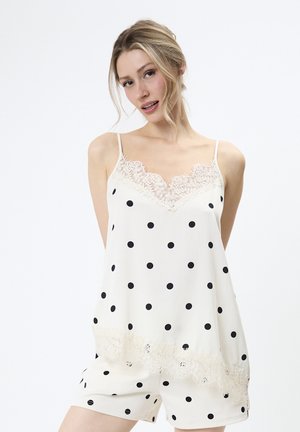 Jonge vrouw met een witte top met stippen en shorts met kanten details, staand tegen een effen witte achtergrond.