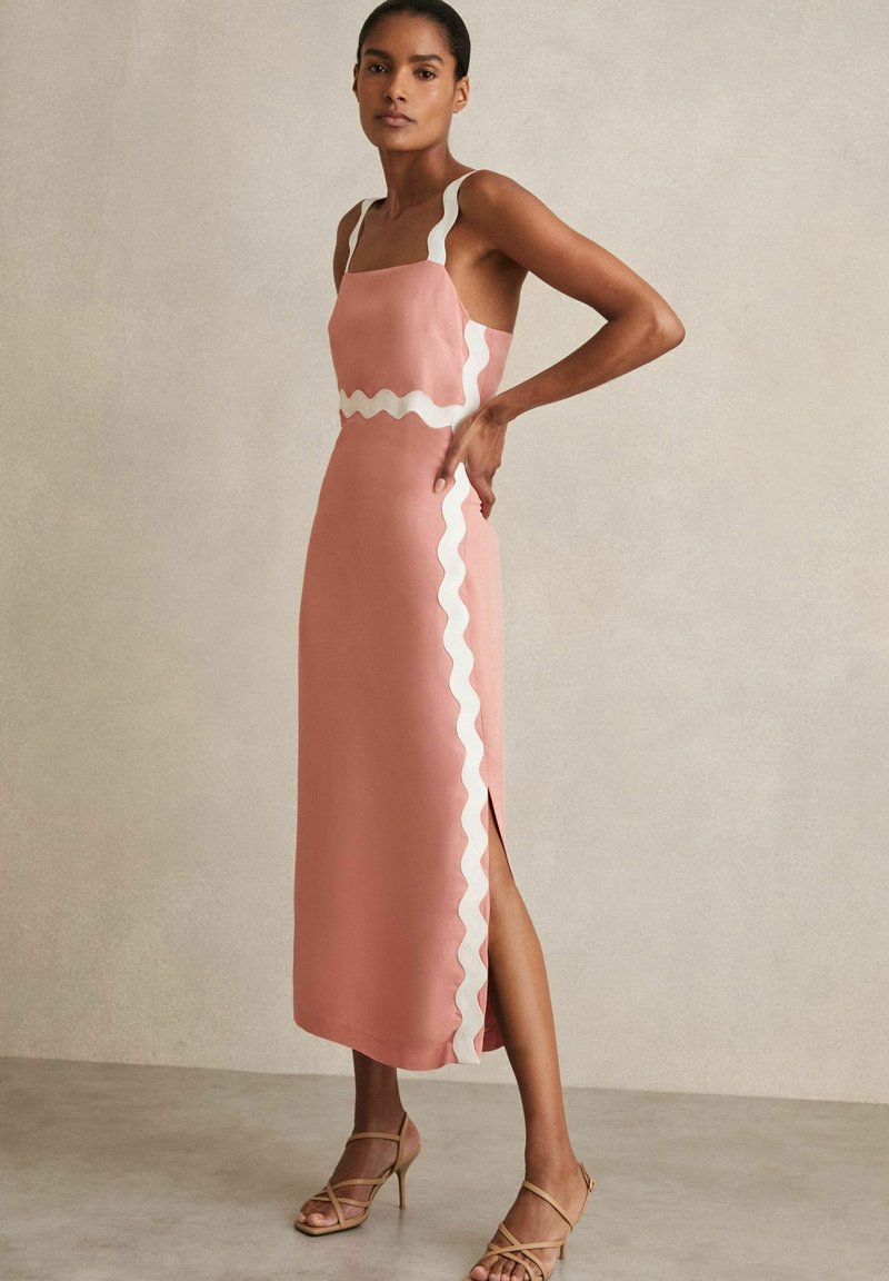 Rosa Midi-Kleid mit wellenförmigem weißen Saum, tailliertem Oberteil und seitlichem Schlitz. Der glatte Stoff und die dünnen Träger unterstreichen das Design. High Heels vervollständigen den Look.