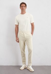 Crème t-shirt met korte mouwen en geborduurd logo, gecombineerd met lichtbeige sweatpants met zijnaad, en witte sneakers met een tan zool.