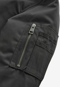 Manche en tissu noir avec une poche à fermeture éclair argentée, comprenant un panneau vertical supplémentaire avec plusieurs fentes de rangement. La texture est lisse.