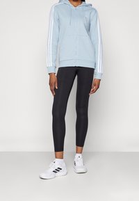 Sudadera azul claro con cremallera y rayas blancas en las mangas, combinada con leggings negros y zapatillas blancas con acentos negros.