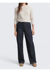 Femme portant une chemise beige clair à manches longues, un jean large et foncé, et des chaussures beiges, debout devant un fond clair uni.