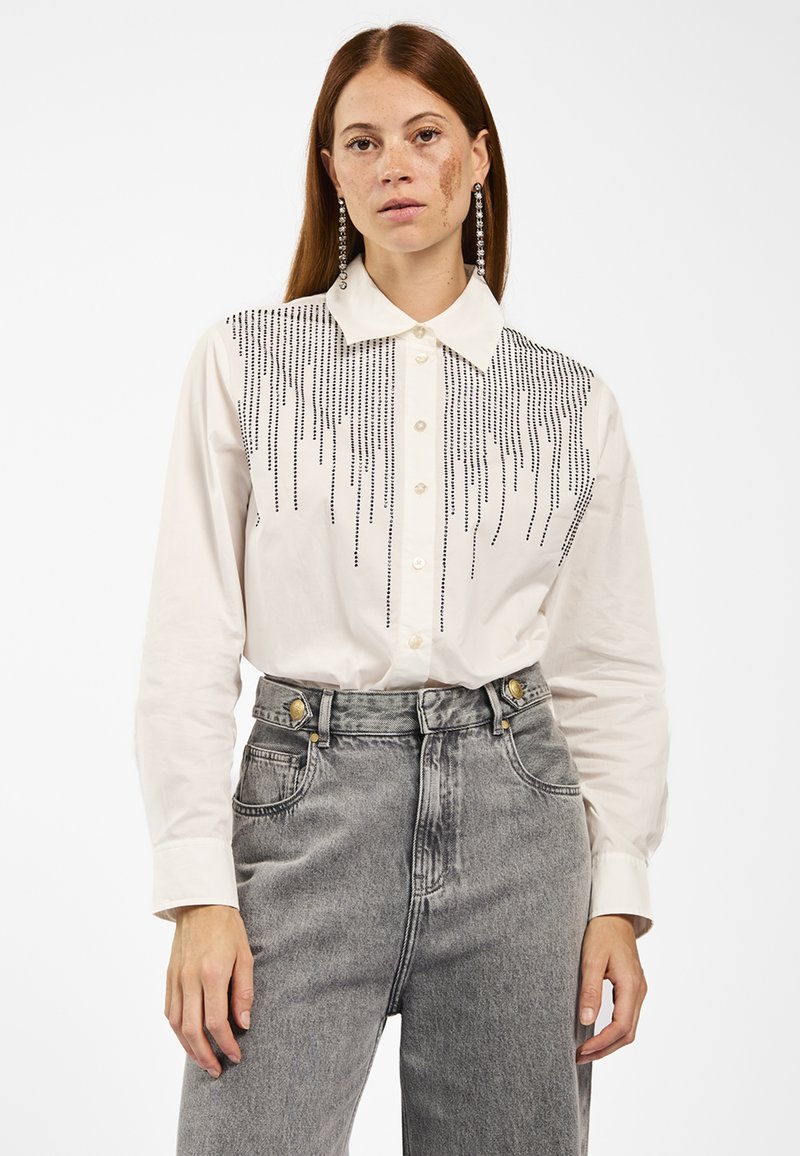 Weißes Langarmhemd mit schwarzen vertikalen Perlenverzierungen auf der Brust, kombiniert mit hoch taillierten grauen Jeans.