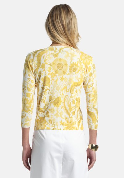 Femme aux cheveux blonds portant un haut à manches longues à motif floral jaune et un pantalon blanc, vue de dos, avec un bracelet manchette en or.